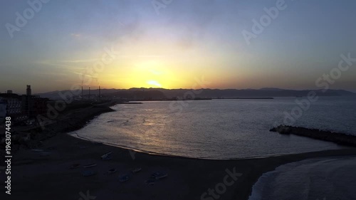 Aerial orbital movement over a tranquil Mediterranean beach and stone breakwater at sunrise, capturing the calm waves and golden horizon in Algeria.