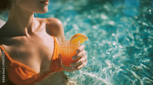 High angle portrait of a person in an orange bikini holding a tropical cocktail by a pool with sparkling turquoise water.