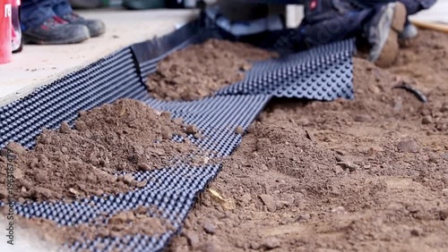 Worker laying down drainage membrane on soil surface, with dirt piles and construction tools visible in the background, demonstrating a methodical installation process