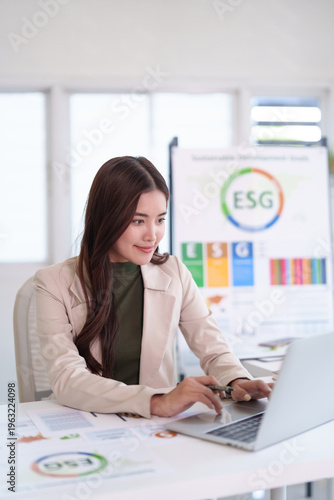 Asian adult businesswoman engaged using laptop analyzing ESG sustainable development goals corporate strategy report at bright modern office environment