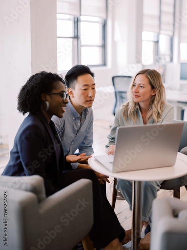 Three professionals engaged in discussion around a laptop at a modern office workspace, featuring a black woman, an Asian man, and a Caucasian woman in business attire