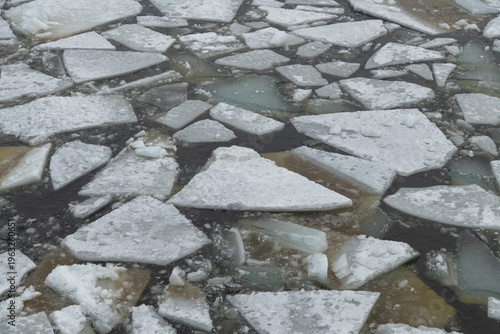 Frozen ice is breaking apart in the water, creating a pattern of fragmented shapes. The scene shows natural ice formations during the winter time. The water moves around the pieces.