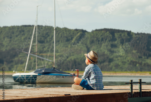 pensive young boy wearing straw hat and plaid shirt sitting on wooden pier looking at sailboat on calm lake as concept of childhood dreams, peaceful solitude and summer vacation in countryside