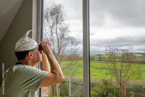 Tinfoil hat picture. A man wearing a tinfoil hat looks through a pair of binoculars towards the sky. Paranoia and conspiracy theory. Alien abduction and government cover up. Mind control and brainwash