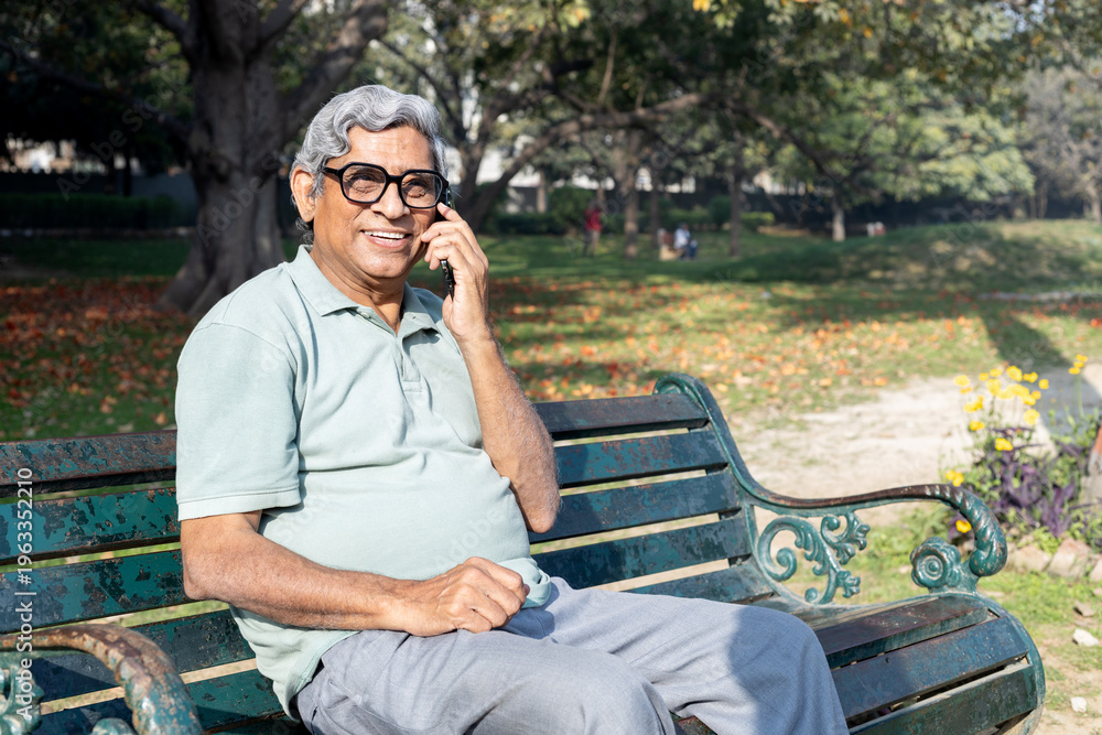 Fototapeta premium Senior Indian man talking on smartphone while sitting on a park bench. Concept of communication, connection, and modern elderly lifestyle.