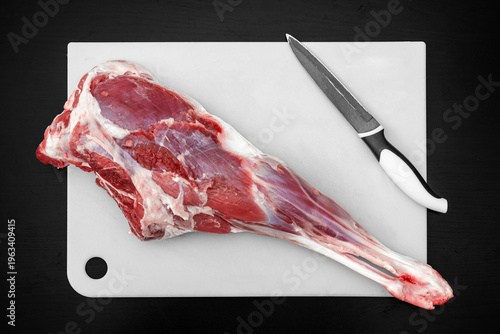 A large piece of raw meat rests on a white cutting board in a kitchen. A sharp knife lies next to the meat, ready for cutting. The kitchen has simple lighting and clean surfaces