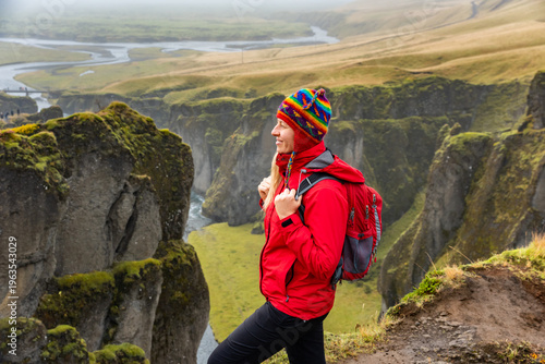 Smiling hiker with a red jacket and colorful hat stands at the edge of Iceland's Fjadrargljufur Canyon, taking in the breathtaking landscape of steep cliffs and winding river below