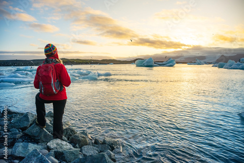 Woman in red jacket joyfully poses on rocky shore of a glacier lake in Iceland. Smiling broadly, she spreads her arms wide, capturing the beauty of the glowing sunset