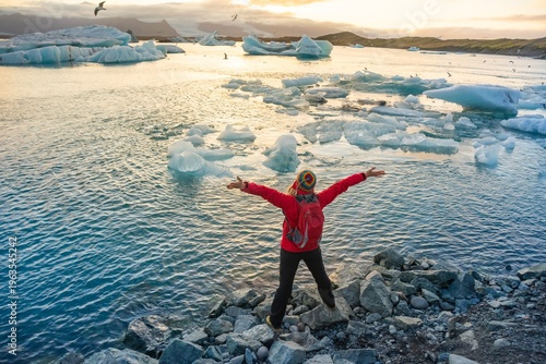 A person stands with arms raised by the ice lagoon in Iceland, celebrating the natural beauty at sunset. Icebergs float in the water, with a colorful sky overhead.