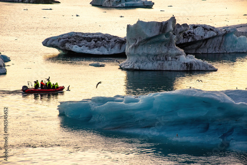 A group of tourists on a boat navigates through a glacier lagoon filled with icebergs. The golden hues of sunset reflect on the water, creating a stunning backdrop