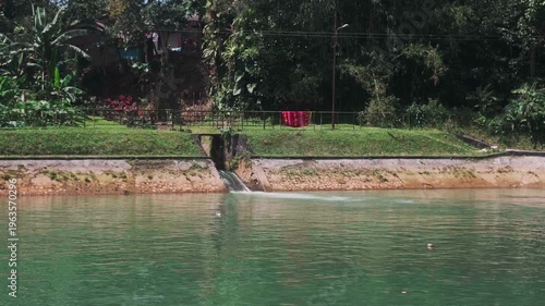A cinematic wide shot of a concrete river dam designed for hydroelectric power generation. The footage captures the steady flow of water through an open spillway, merging with a calm, greenish reservo