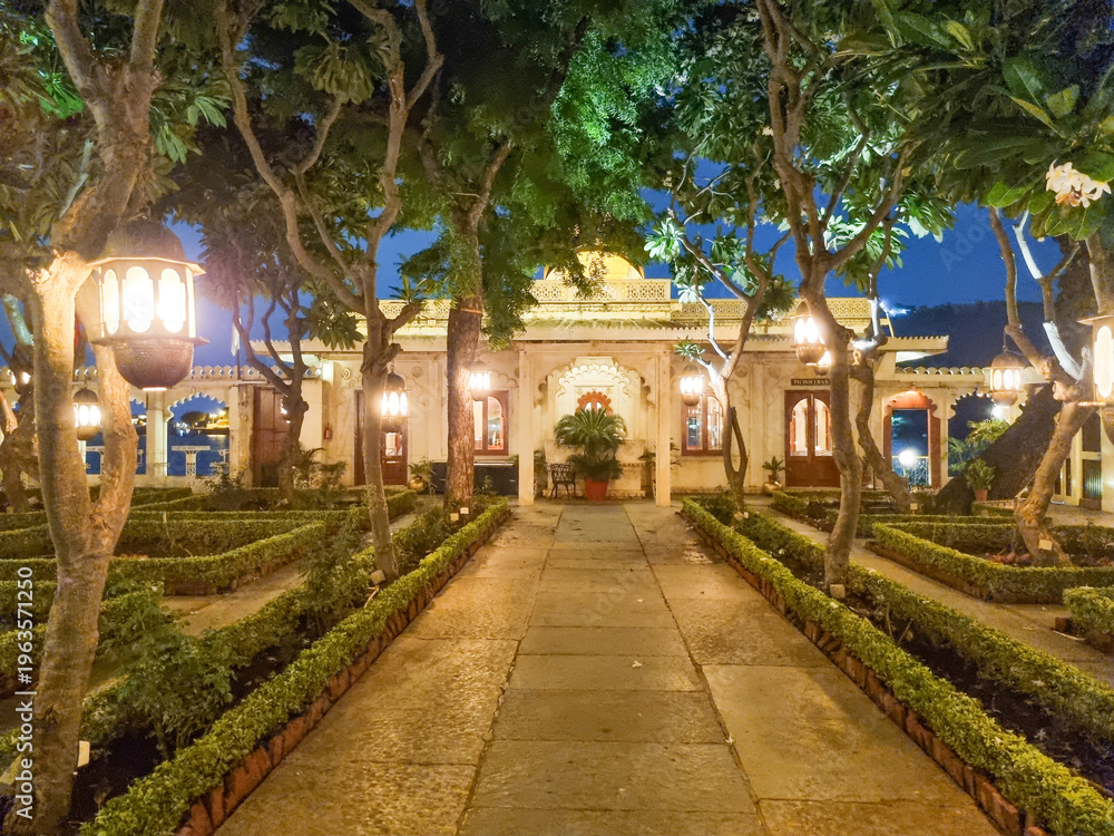 Obraz premium Beautiful night view of Jagmandir Island Palace in Udaipur with glowing lights, symmetrical garden pathway, trees, and heritage architecture reflecting royal ambiance in India