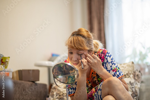 Middle aged woman applying eyeliner looking at small mirror in living room. Daily morning beauty makeup routine of 57 year old lady. Self care and aging gracefully concept at home.