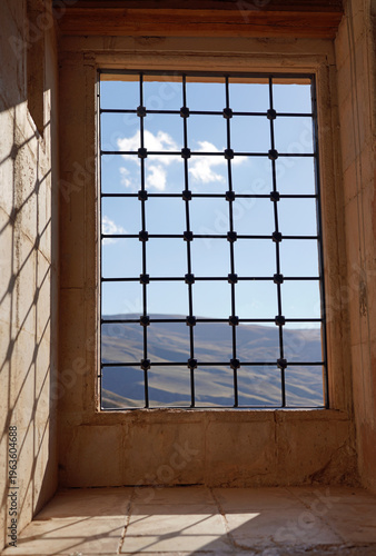A view of the rolling Anatolian hills through an iron-grilled window of the historic Ishak Pasha Palace, casting geometric shadows across the ancient stone interior
