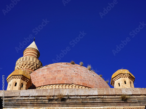 A low-angle view of the weathered stone domes and ornate minaret of the historic Ishak Pasha Palace set against a clear, deep blue sky in Doğubeyazıt, Turkey