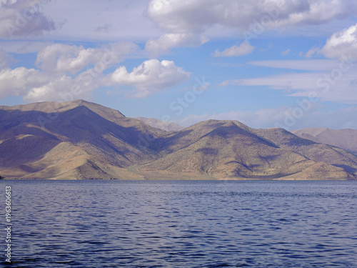 The vast, shimmering blue waters of Lake Van meet the rolling, arid mountains of Eastern Anatolia under a bright sky with scattered white clouds