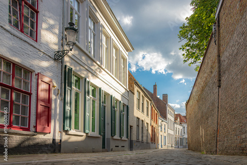 Traditional Flemish brick houses along a quiet cobblestone street in Bruges, Belgium. Historic medieval architecture in the UNESCO World Heritage city center on a clear day.