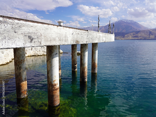 A weathered concrete pier supported by cylindrical pillars extends over the clear turquoise waters of Lake Van on Akdamar Island, with a majestic mountain peak visible in the distance