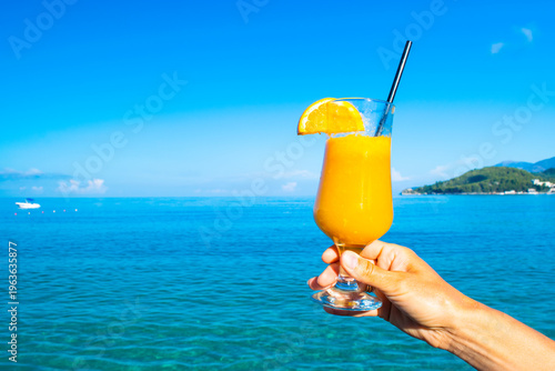 Woman's hand holds a glass of orange fresh juice with a straw and an orange slice against the background of a blue sky and a calm sea.