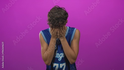 Teen boy in a blue basketball jersey covers his face with hands and rubs his eyes in a studio; sadness vulnerability.