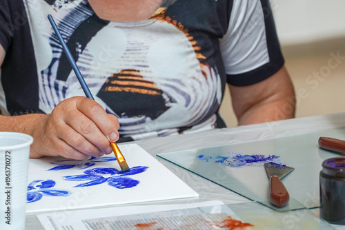 Woman painting blue floral shapes on paper using brush during art workshop. Focus on creative expression, artistic hobby and calm handmade activity in indoor studio setting