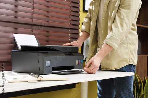 Foto Man taking printed sheet of paper from printer at white desk indoors, closeup