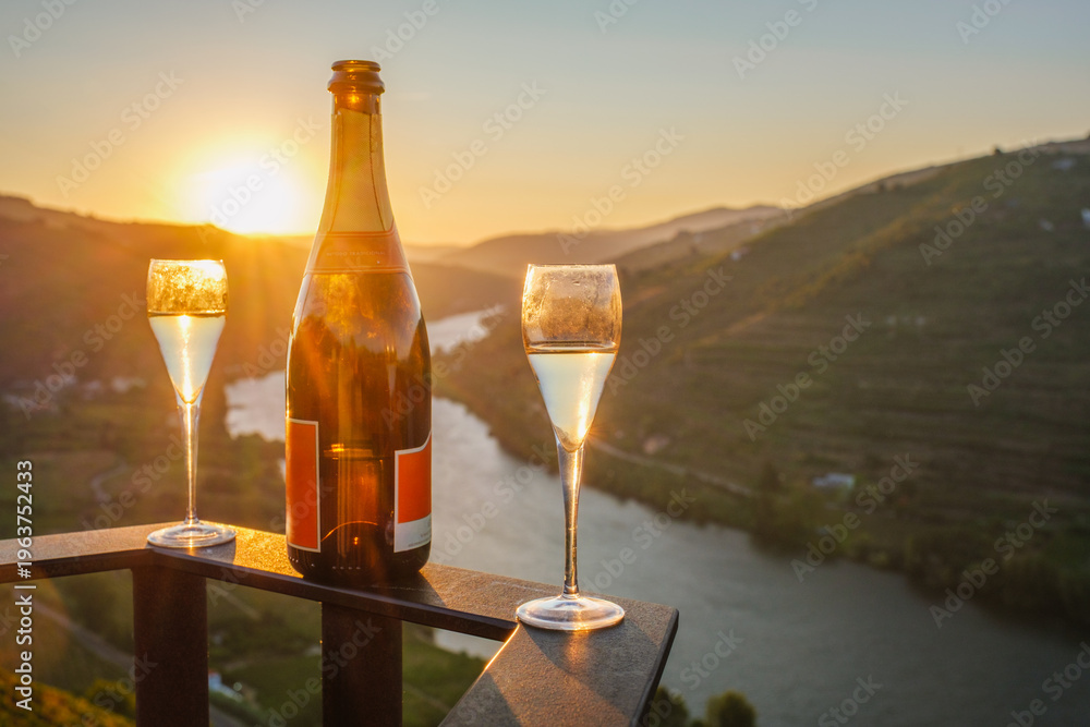 Naklejka premium Bottle of sparkling wine with two filled glasses overlooking the Douro River and vineyard hills at sunrise in the Douro Valley, Portugal.