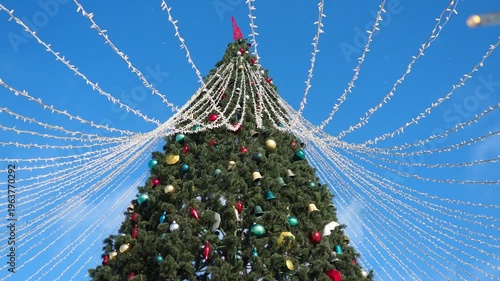 A grand tree shines brightly, adorned with colorful ornaments and sparkling lights. Against the clear blue sky, the holiday spirit comes to life, wrapping the world in joy and warmth
