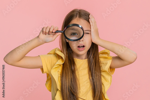 Young child girl looks through a magnifying glass and peers into camera with a joyful smile, pretending to investigate or explore. School kid isolated on pink background with curious playful emotion.