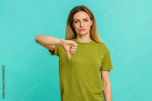 Middle-aged woman gives thumbs down with firm expression, showing disapproval, dissatisfaction, rejection of idea message or result. Girl isolated on blue background displays negative response clearly