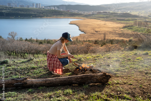 Canvas Print Serene young woman traveler sitting near small campfire in nature by lake and hills enjoy calm time