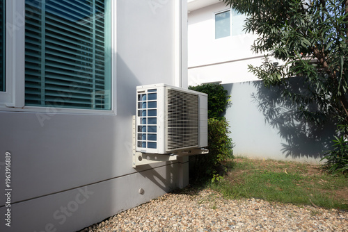 Outdoor air conditioning unit on side of residential house, system mounted on exterior wall, positioned for efficient airflow and effective heat exchange, ensuring comfortable indoor conditions.