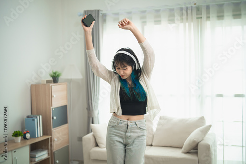 Young woman enjoying music at home, dancing with headphones on, raising hands in joy, casual attire, vibrant hair, cozy living room setting with soft light