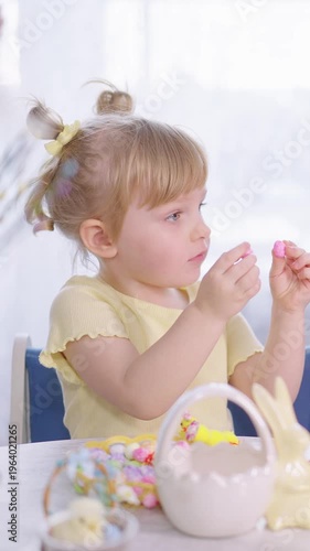 Little girl sits at table with Easter basket. She has pigtails and wears yellow dress. Hands reach for colorful candies with focused eyes. Bright daylight illuminates the cheerful scene