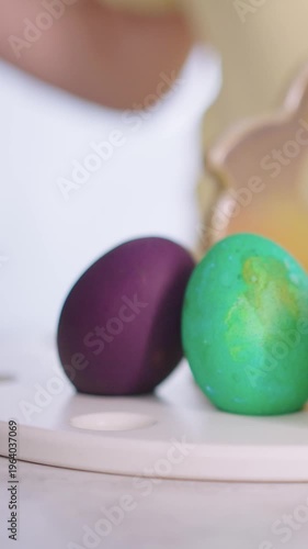 Soft hand gently touches colorful Easter eggs. Eggs rest on a simple white plate. Child's fingers explore the smooth, dyed surfaces. Joyful, innocent play evokes springtime celebration