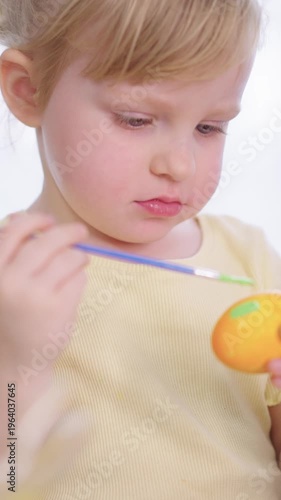 Child paints orange egg with care and focus. Soft light highlights gentle concentration. Green brush strokes add color to the egg. Bright yellow shirt contrasts with calm background