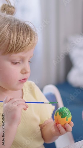 Child paints Easter egg with focused attention. Bright colors fill the egg as brush strokes appear. Soft light enhances calm, creative atmosphere. Perfect for spring, family, holiday