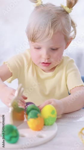 Tiny girl with pigtails focuses on colorful eggs. She uses wooden tools with care and concentration. Her small hands move slowly, showing quiet attention. Soft light highlights her calm