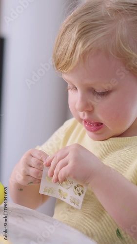Child focuses on a sticker with curiosity and quiet concentration. Soft light highlights baby's gentle hands and thoughtful expression. Table holds a green apple beside the sticker