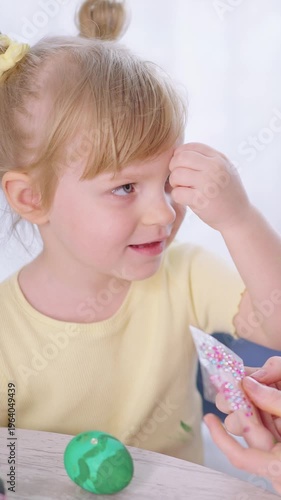 Little girl smiles while decorating a green ball with pink sprinkles. She wears a yellow shirt and has blonde hair in a bun. Her hands gently sprinkle glitter over the ball