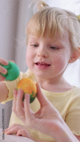 Sunlit room. Little girl smiles. Hands hold colorful eggs. Child's eyes bright with delight. Joyful play. Soft daylight filters through window. Perfect for Easter scenes. Child wears yellow top