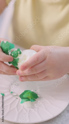 Child holds green egg gently, showing care. Tiny hands paint egg, creating bright patterns. Plate holds paint splatters, hinting at playful creativity. Soft yellow shirt adds warmth to the scene