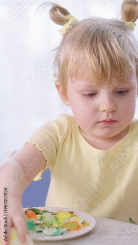 Tiny girl holds egg, focused, curious. She wears yellow shirt, pigtails tied with clips. Child's hands carefully peel eggshell with care. Plate holds colorful Easter eggs beside her