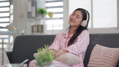 Asian adult woman peacefully listen music headphones comfortable sofa enjoying quiet time living room home environment