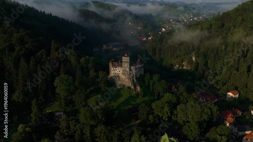 Iconic Bran Castle illuminated by bright summer sunrise over forested hills with mist, high-altitude straight forward aerial drone flight graded