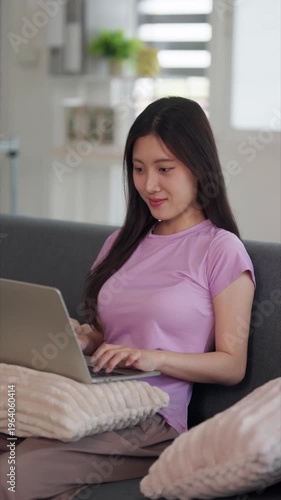 Asian adult woman uses laptop computer remote work online study browsing internet home comfortable sofa living room modern apartment digital lifestyle