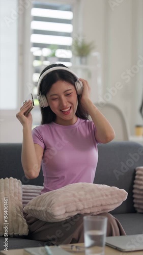 Asian adult woman listening music headphones smartphone smiling enjoying online entertainment home living room relaxed leisure digital media technology