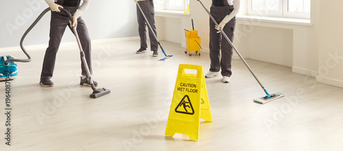 Cleaners team janitors mopping vacuuming sanitation safety. They maintain hygiene in a bright workplace with mops and a vacuum while a wet floor sign warns for safety. Clean team at a job.