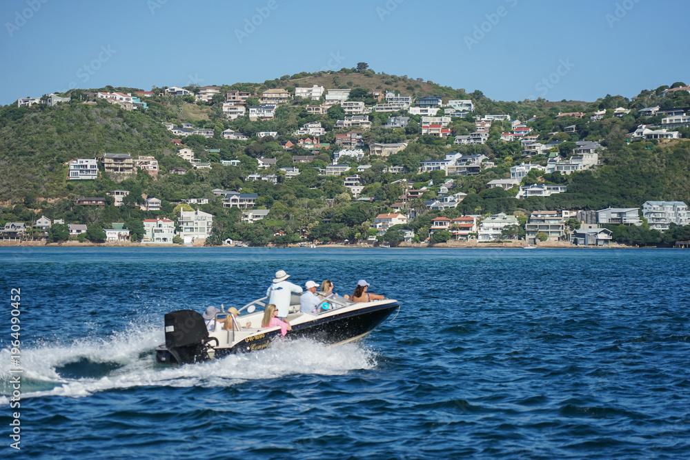 Fototapeta premium South Africa,Knysna Lagoon,A speedboat carrying passengers cruises across calm blue waters near a hillside town with houses scattered across the slope.