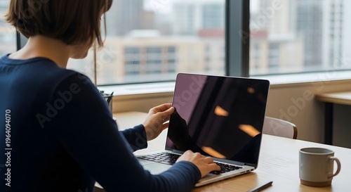 Woman typing on laptop at desk with coffee and city view with over the shoulder